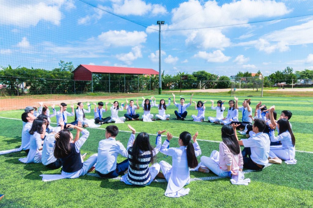 people-sitting-on-green-lawn-grass-while-doing-hands-up-at-daytime-1164572 Students seated in a circle on a school field engaging in outdoor activities.